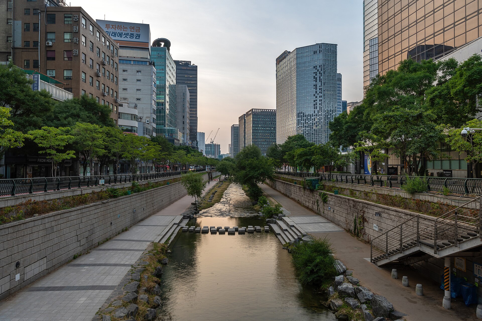 Suối Cheonggyecheon chảy giữa hai bờ cây xanh và lối đi bộ, cao ốc Seoul hai bên, đá bước qua suối ở giữa
