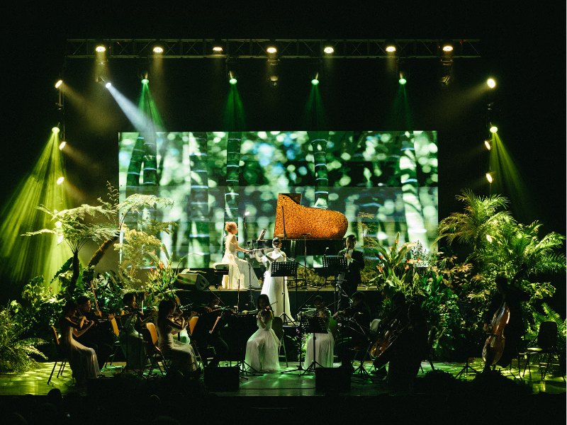 Sân khấu hòa nhạc Voice of Nature 2025 tại Nhà hát Bến Thành: hai cánh sân khấu phủ dương xỉ thân gỗ và chuối cảnh, dàn nhạc chơi giữa mảng cây