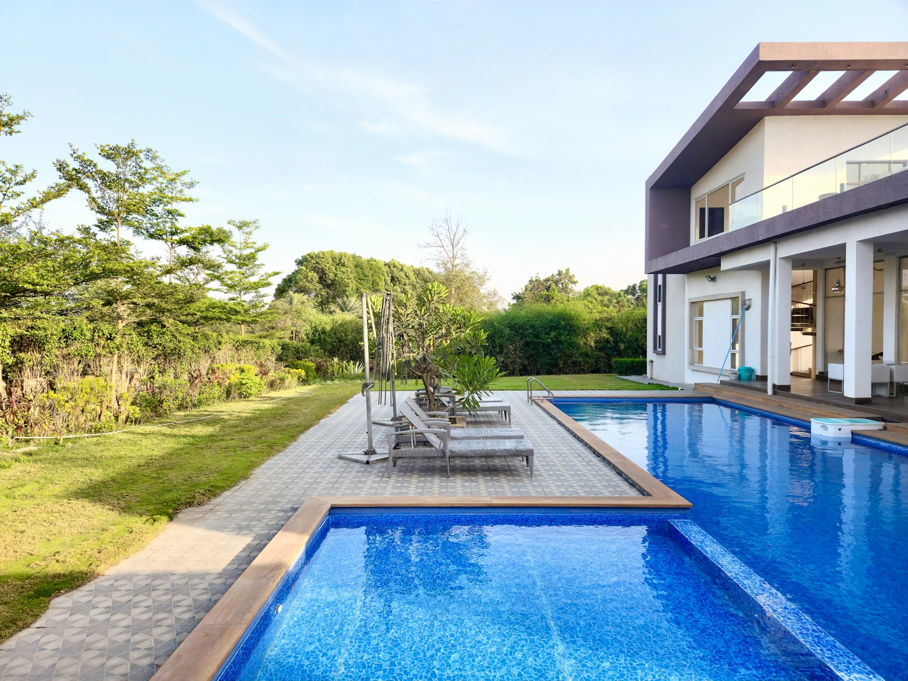 Bể bơi chữ nhật viền sàn lát cạnh khối nhà trắng nhiều cửa, bãi cỏ và cây xanh bên trái