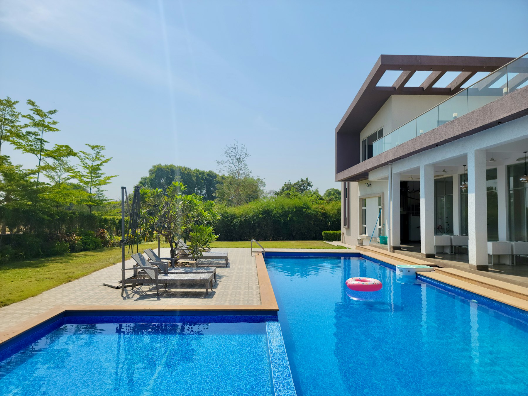 Bể bơi hình chữ nhật cạnh khối nhà trắng, ghế nằm trên sàn lát, quả bóng nổi trên mặt nước, bãi cỏ và cây phía sau