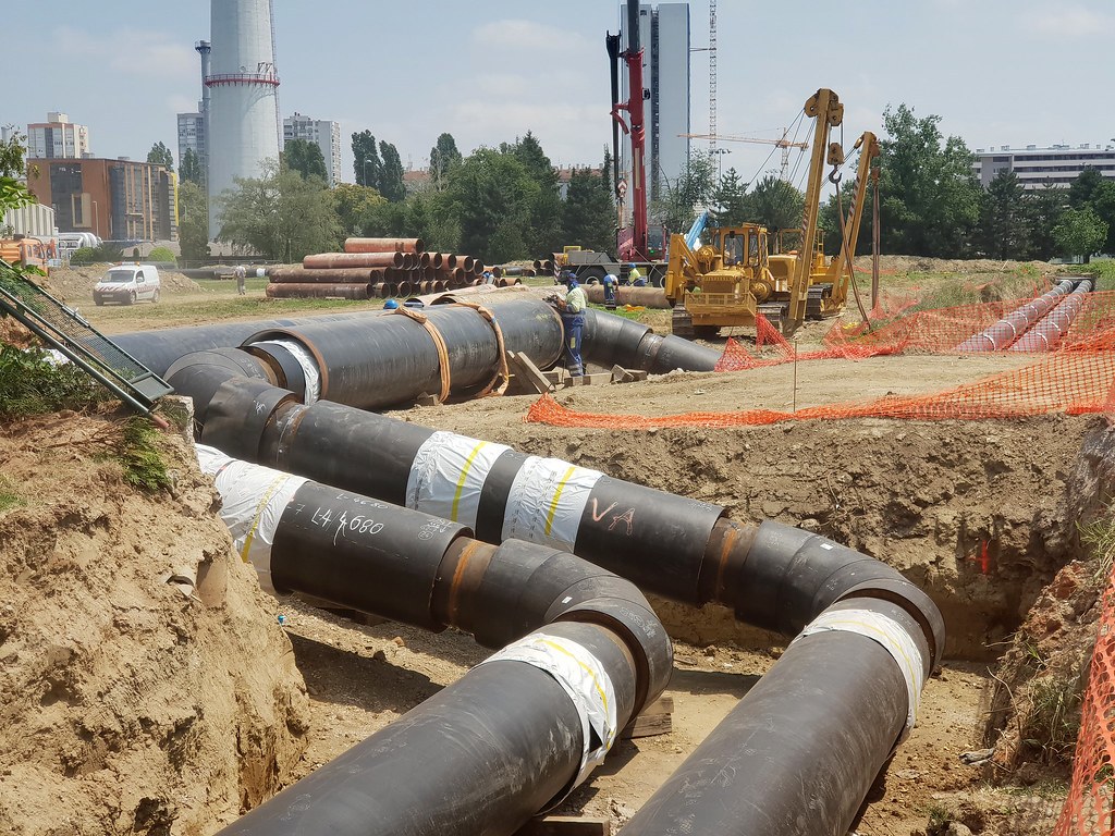 Công trường đặt ống sưởi bọc nhựa đen trong một hào đất đã đào sẵn, cần cẩu vàng và xe tải phía xa