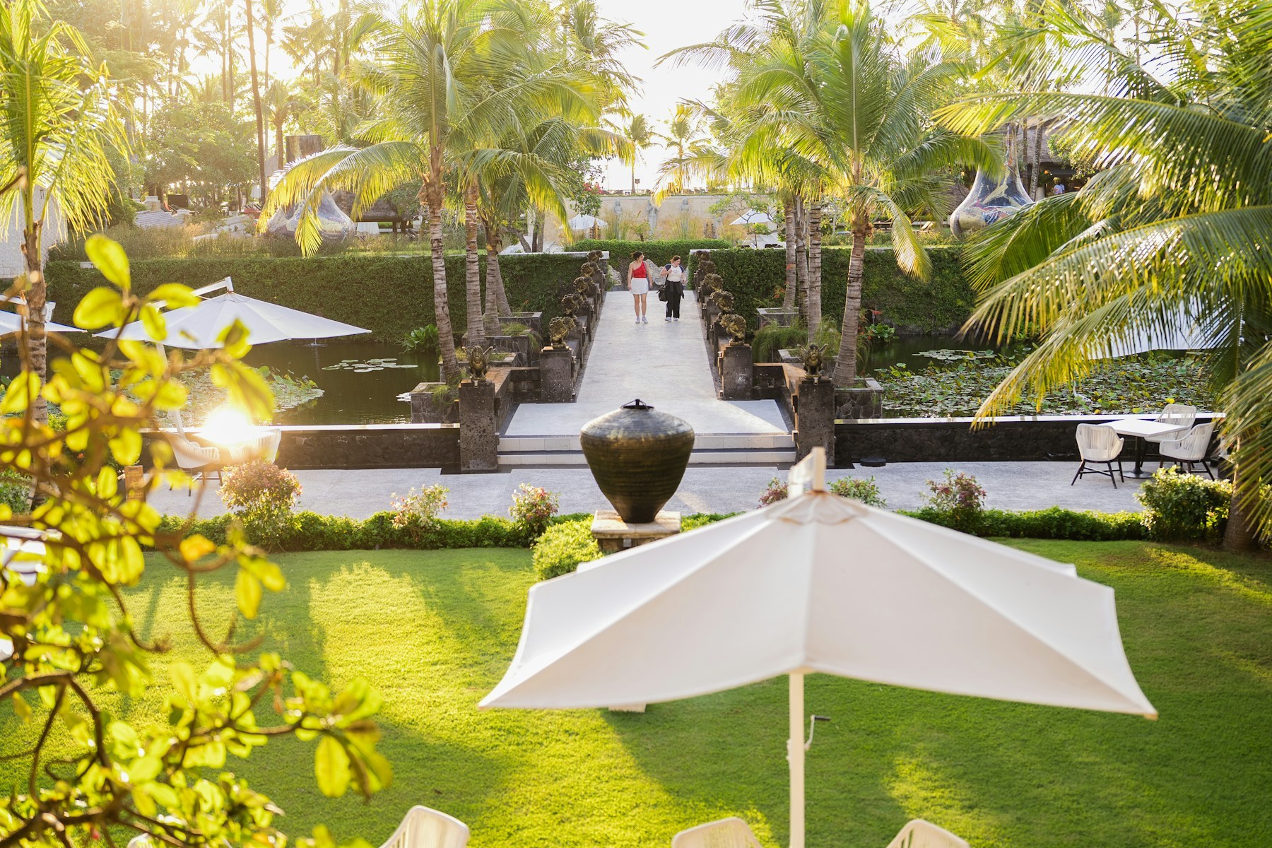 Lối lát đá bắc qua ao sen, hai hàng cột đá kiểu Bali, hai người đi bộ ở xa, dừa và hàng rào cây xén