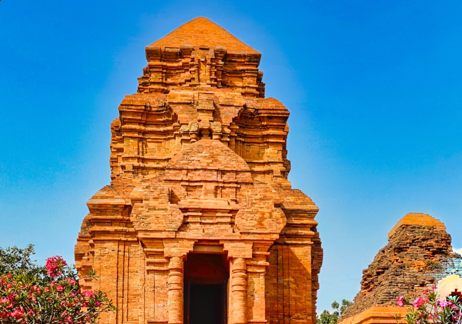Tháp Champa nhìn cận từ phía trước, cửa chính hình vòm cuốn nhô ra khỏi mặt tường, có bụi cây hoa trước sân
