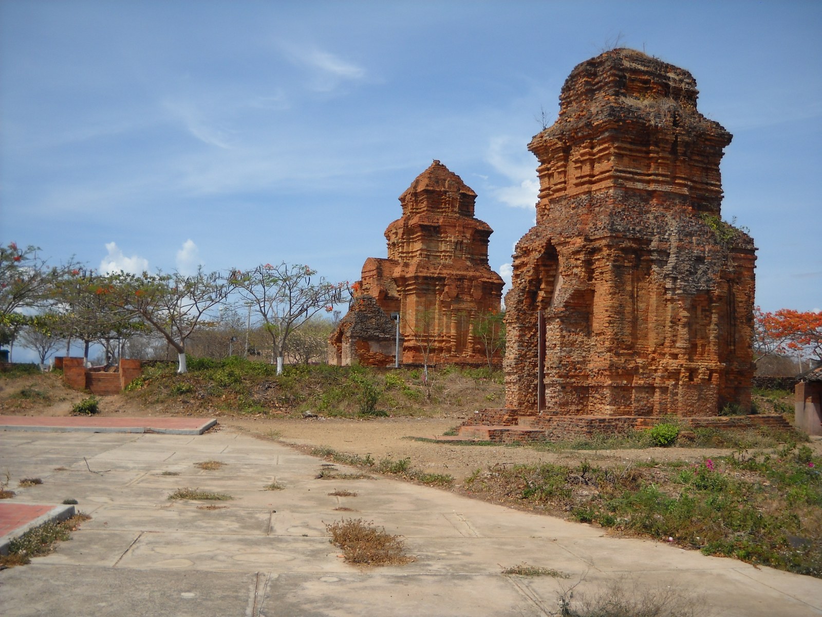 Ba khối tháp gạch Champa đứng trên một khoảng sân đất trống, nhìn từ xa dưới nền trời nhiều mây