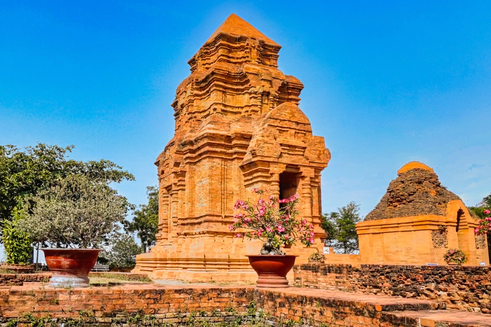 Một tháp Champa màu gạch đỏ nhìn từ sân trước, phía dưới có chậu cây cảnh và bệ đá nhỏ