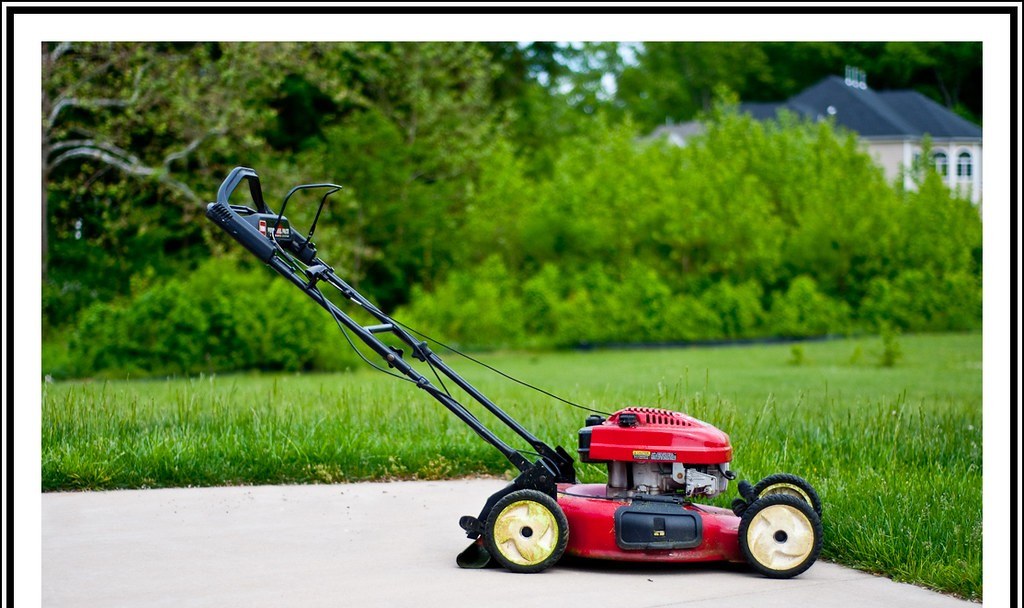 Máy cắt cỏ đẩy tay màu đỏ đỗ trên lối bê tông cạnh bãi cỏ và hàng cây