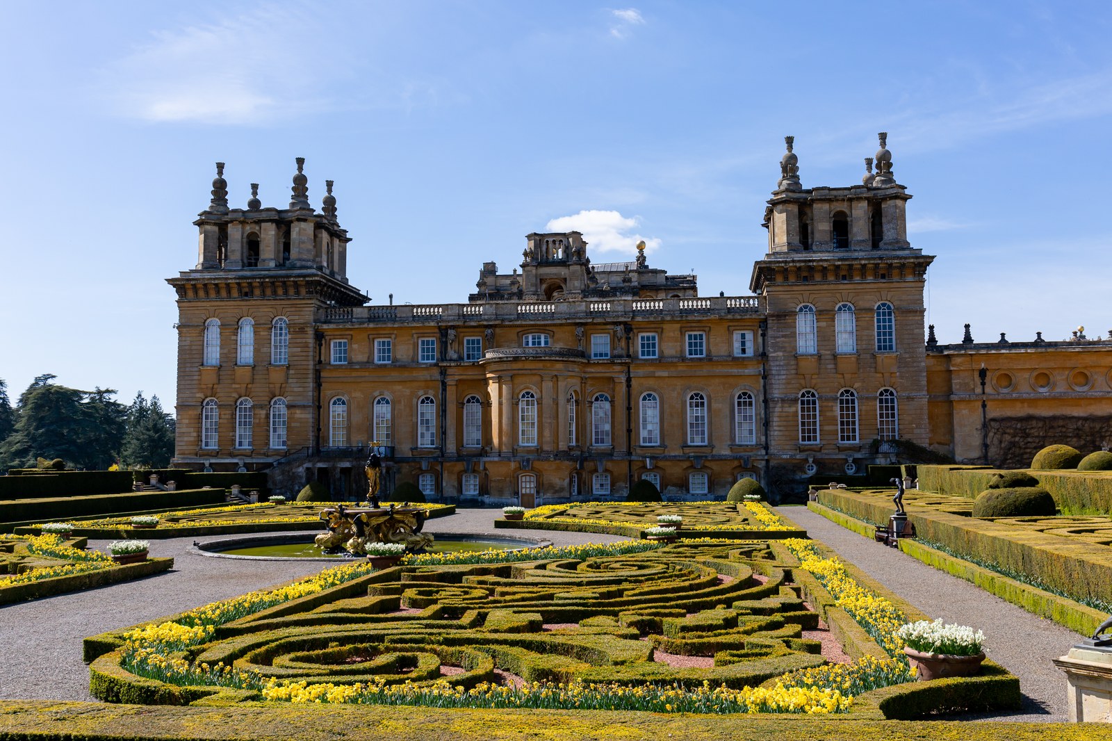Mặt tiền cung điện đá lớn kiểu Baroque, phía trước là vườn parterre hình cuộn với hàng cây bụi cắt xén thấp, hoa thuỷ tiên vàng, và một đài phun tượng vàng giữa bồn nước tròn.