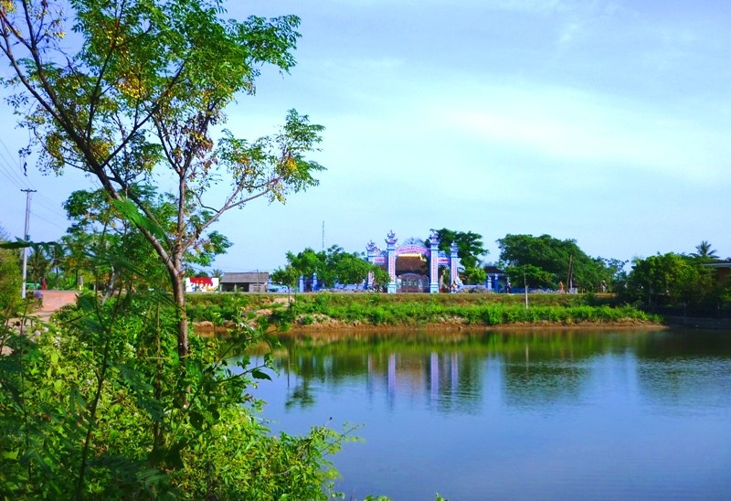 Cổng đình Mai Xá nhìn qua một ao nước lớn, cổng sơn xanh tím có mái nhỏ, cây cối bao quanh bờ ao