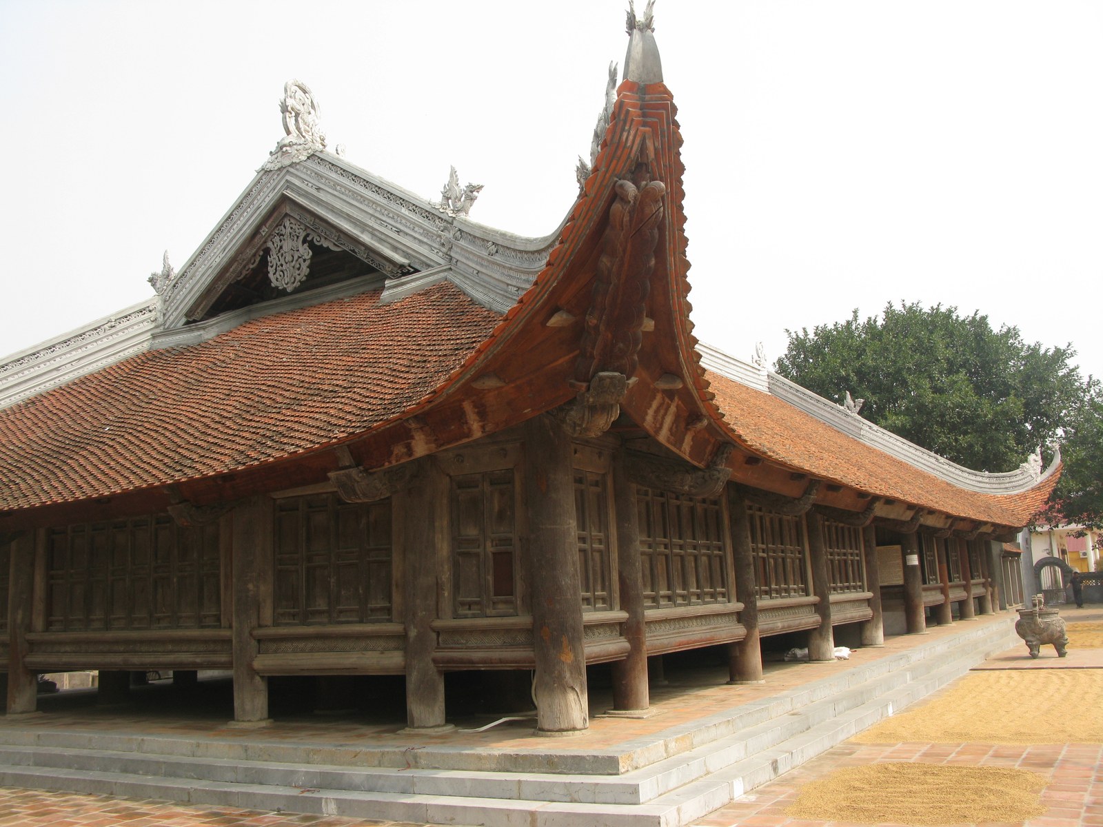 Toàn cảnh đình Đình Bảng (Bắc Ninh) nhìn chéo: mái ngói đỏ cong hai tầng, hàng cột gỗ tròn đỡ hiên rộng, sân gạch phía trước có thóc phơi
