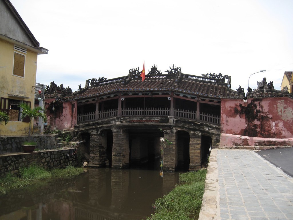 Chùa Cầu Hội An nhìn từ bờ kênh: mái ngói cong nối hai bờ, các nhịp vòm đá bên dưới, cây xanh và nhà cổ hai bên