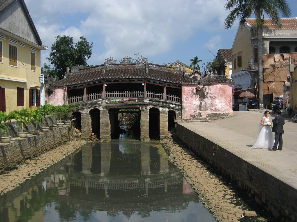 Chùa Cầu Hội An nhìn ban ngày, có hai người mặc áo cưới đứng gần đó, hai bên là nhà cổ và hàng cây dừa