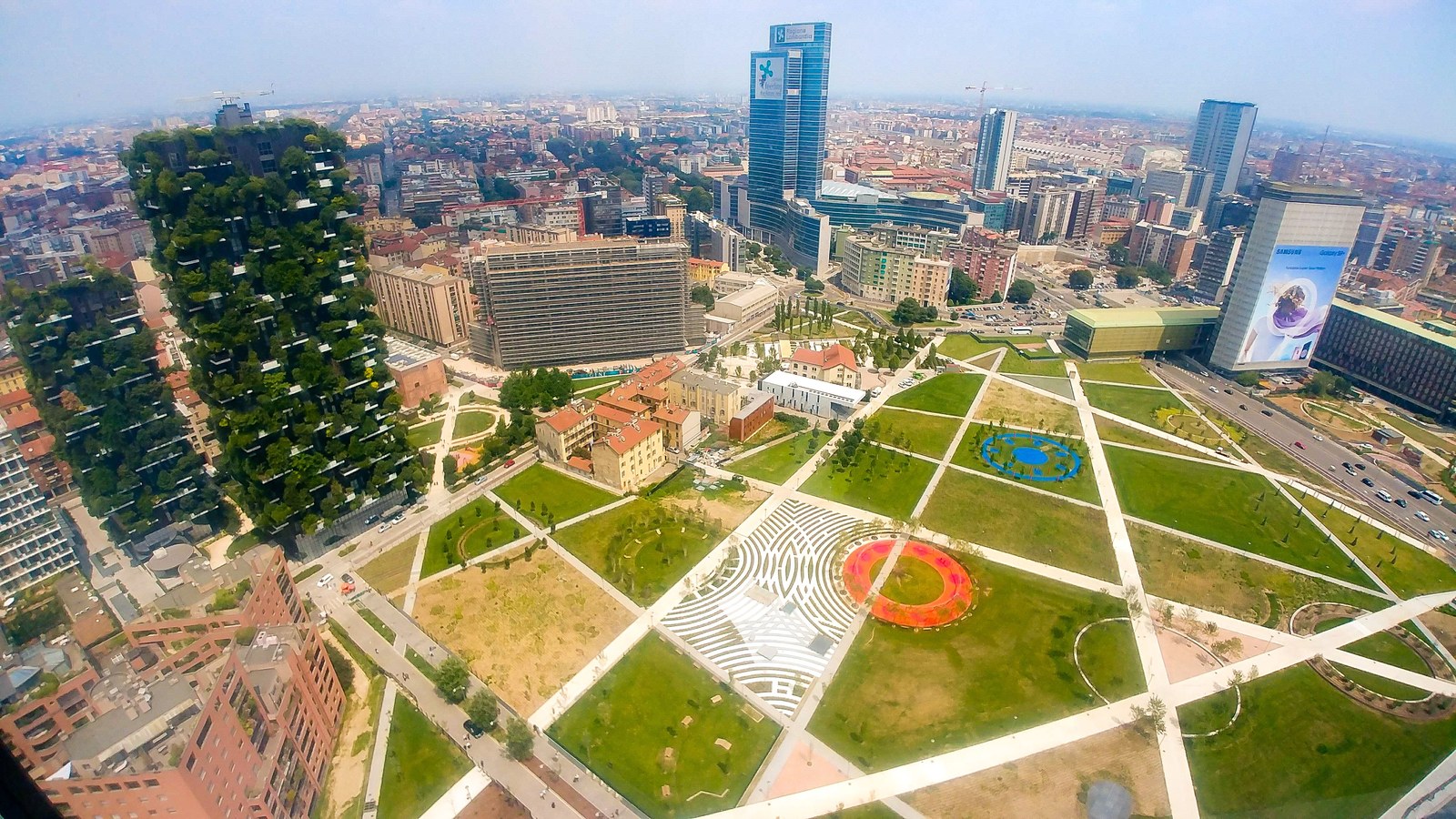 Nhìn từ trên cao xuống công viên Biblioteca degli Alberi ở Milan, hai toà Bosco Verticale phủ cây ở góc trái, phía trước là các ô cỏ và lối đi lát đá hình học