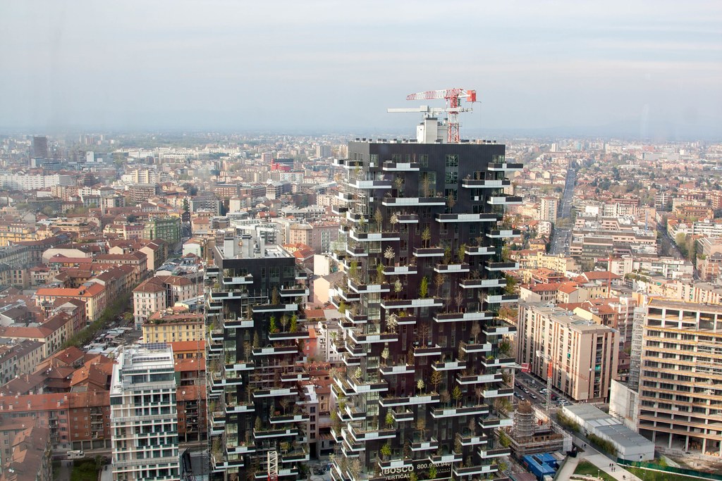 Toà Bosco Verticale nhìn từ góc phố, cạnh một khối nhà màu cam thấp tầng để so sánh tỷ lệ