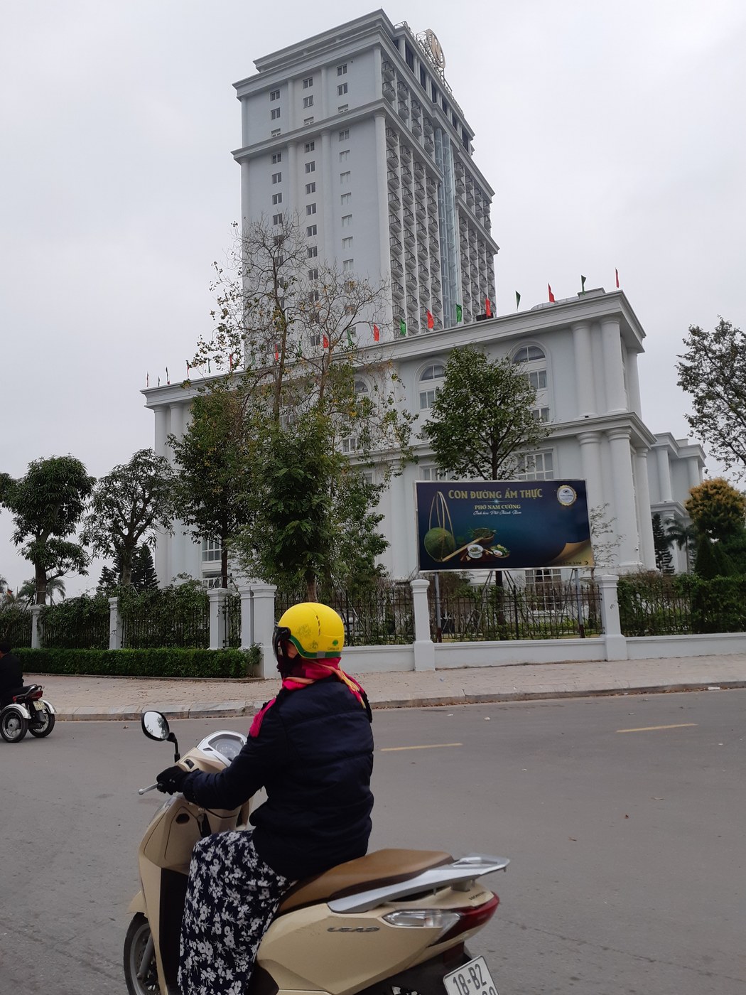 Một khách sạn cao tầng đứng sau hàng rào công trường, người đi xe máy ở phía trước, biển quảng cáo tuyến phố ẩm thực bên đường