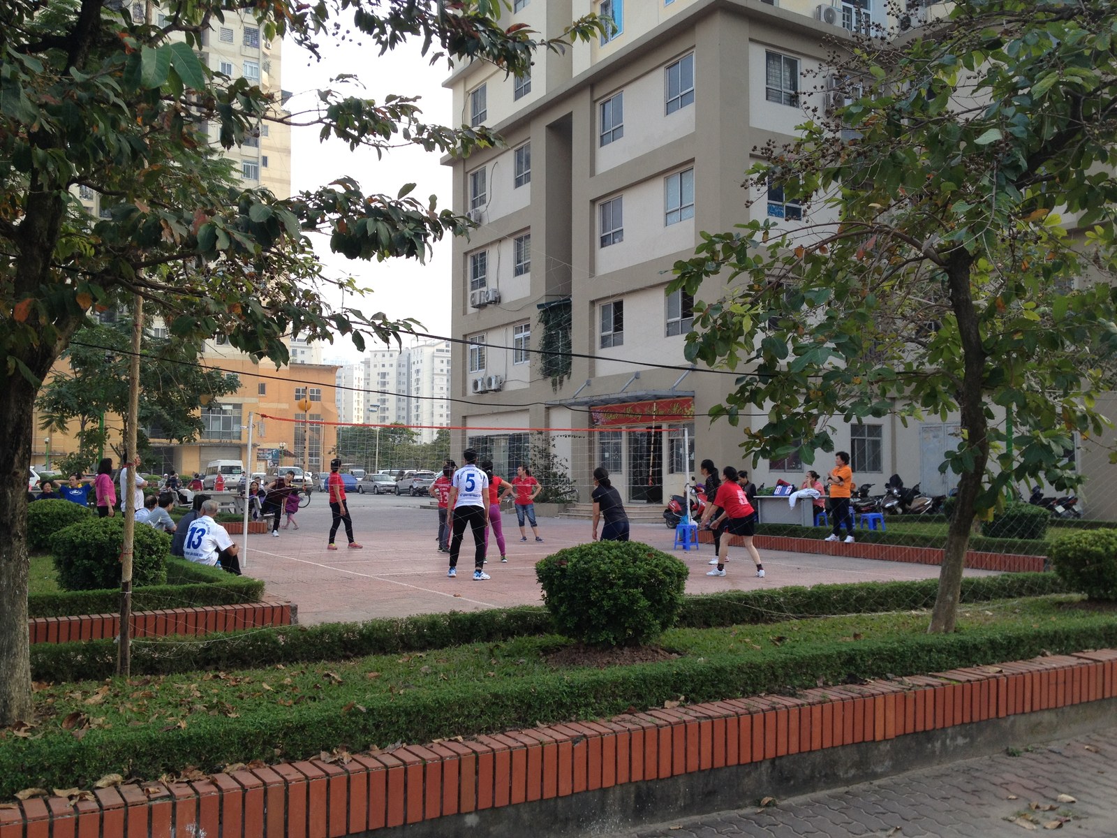 Người dân chơi bóng chuyền trên sân giữa khu chung cư, cây xanh cắt tỉa dạng khối tròn viền quanh sân, các toà nhà cao tầng phía sau