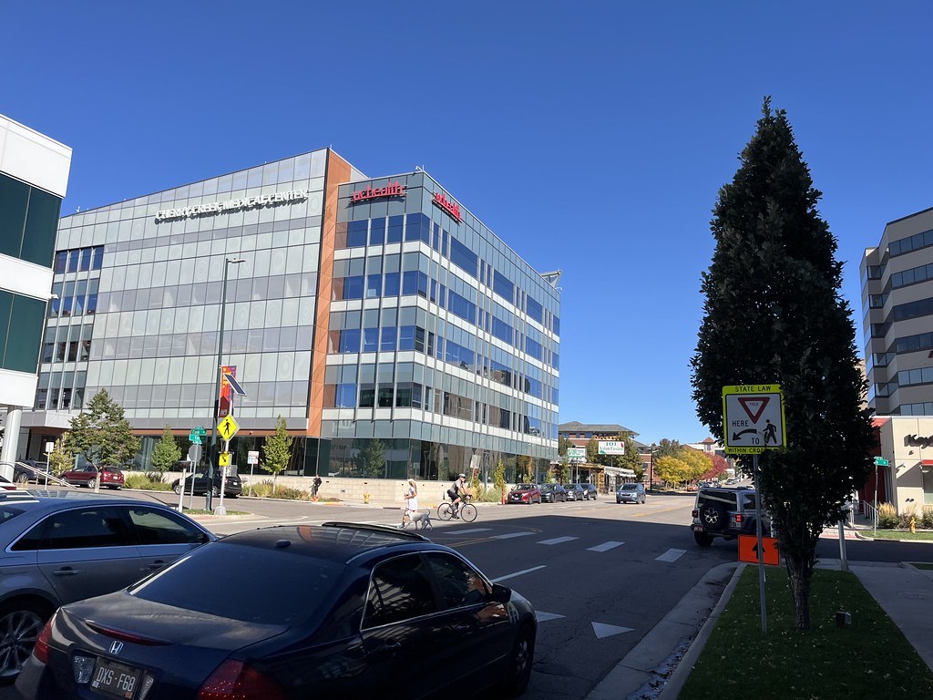 Phố nhiều làn ở Denver: một người đi xe đạp và một người dắt chó băng qua giao lộ trước cao ốc kính, ô tô đậu sát vỉa hè hai bên đường