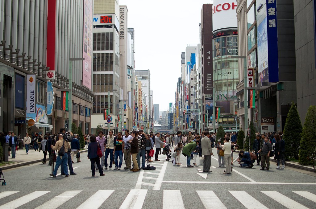 Giao lộ có vạch sang đường kẻ trắng ở khu Ginza, đông người đứng và đi lại giữa lòng đường, hai bên là các toà nhà cao tầng phủ đầy biển hiệu quảng cáo.