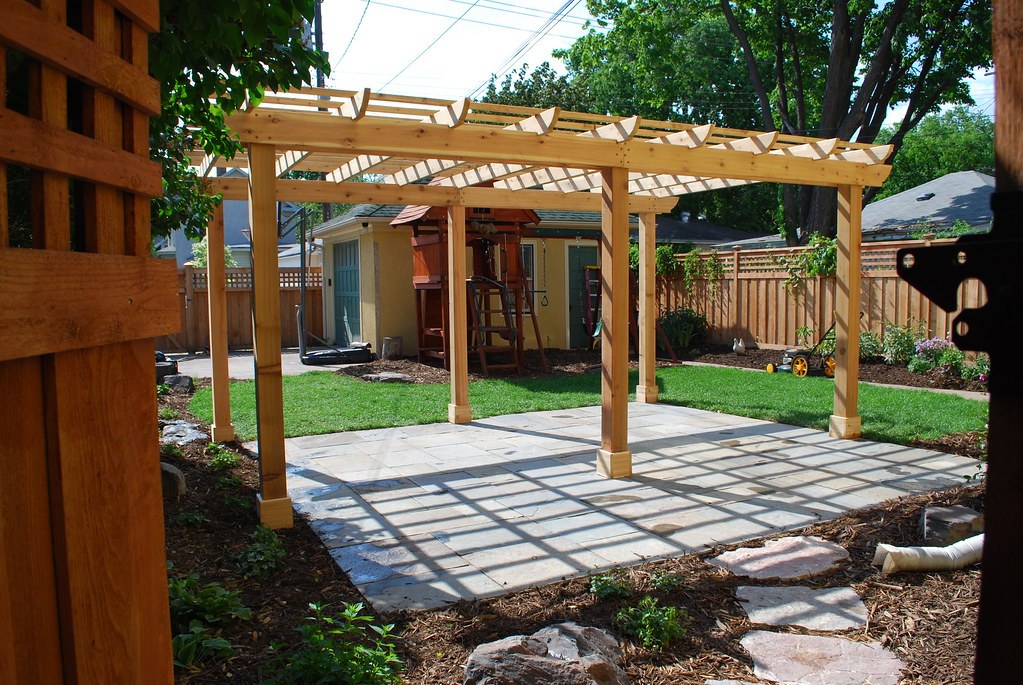 Giàn pergola gỗ để trần thanh ngang, nắng xuyên qua tạo bóng sọc trên sân lát đá, bàn ghế nướng ngoài trời bên dưới